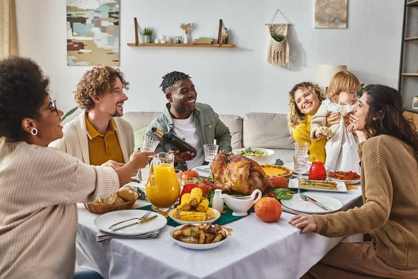 Hombre afroamericano vertiendo vino en el vaso, familia multirracial celebrando Acción de Gracias en casa - foto de stock