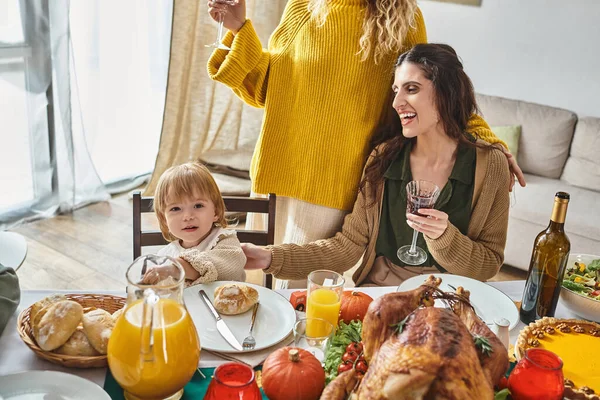Niedliche Kleinkind Mädchen Blick in die Kamera in der Nähe lgbt Eltern und Thanksgiving gebratenen Truthahn auf dem Tisch — Stockfoto