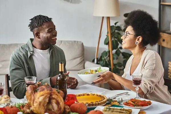 Happy african american woman passing bowl with salad to relative during Thanksgiving dinner, turkey — Stock Photo