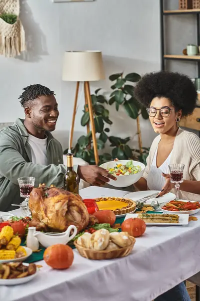 Cheerful african american woman passing bowl with salad to relative during Thanksgiving dinner — Stock Photo