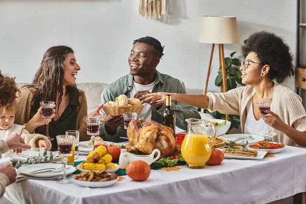 Feliz afro-americana atingindo pães cozidos perto de amigos e familiares durante o jantar de Ação de Graças — Fotografia de Stock