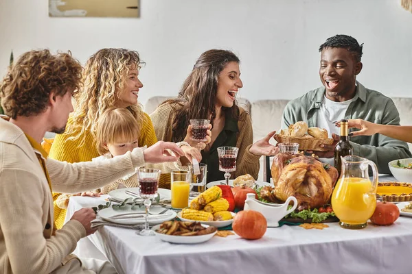 Familia interracial alegre y amigos reuniéndose en la mesa de Acción de Gracias con varias comidas y bebidas - foto de stock