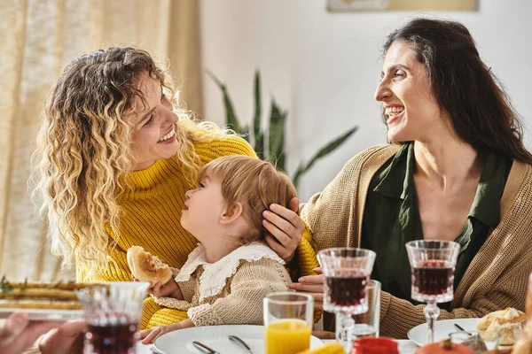 Alegre pareja lesbiana y niño pequeño disfrutando de deliciosa cena mientras se reúnen en Acción de Gracias - foto de stock