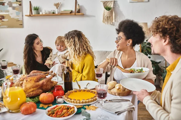 Amigos multiculturales teniendo una deliciosa cena mientras se reúnen en Acción de Gracias, las mujeres calmando a los niños - foto de stock
