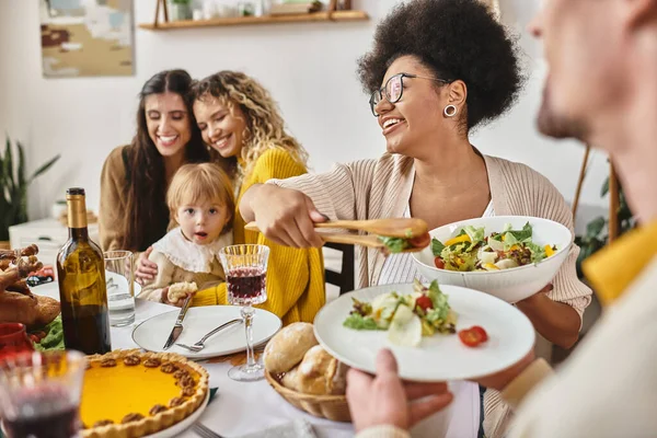 Joyeuse femme afro-américaine servant de la salade à l'homme bouclé le jour de Thanksgiving, famille lgbt et bébé — Photo de stock