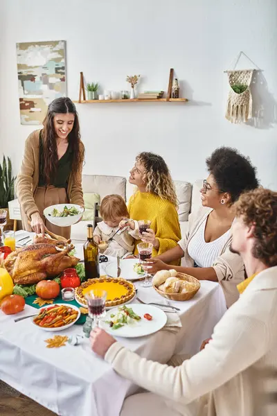Mujer feliz sirviendo comida a amigos interracial y familiares durante la celebración de Acción de Gracias en casa - foto de stock