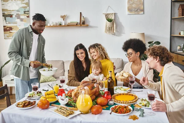 Hombre afroamericano feliz sirviendo comida a amigos interracial y familia en el día de Acción de Gracias - foto de stock