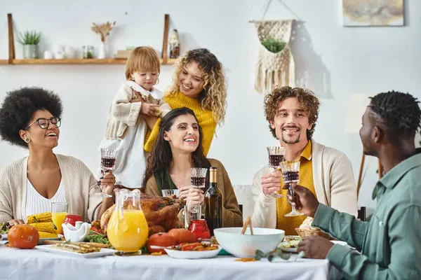 Feliz Día de Acción de Gracias, alegres amigos multiculturales y familia animando con copas de vino tinto - foto de stock