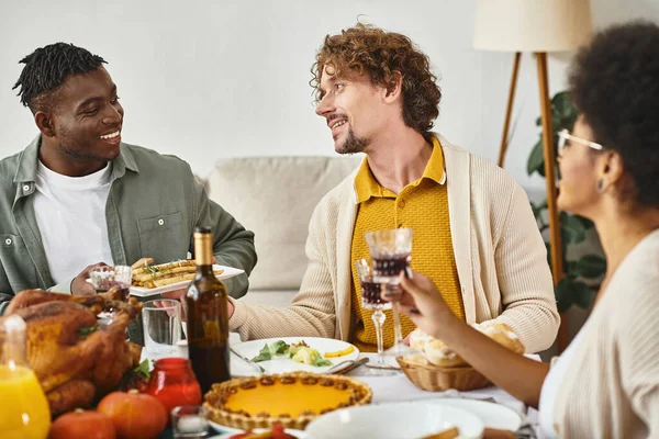 Feliz Día de Acción de Gracias, alegres amigos multiétnicos charlando en la mesa con pavo y pastel de calabaza - foto de stock