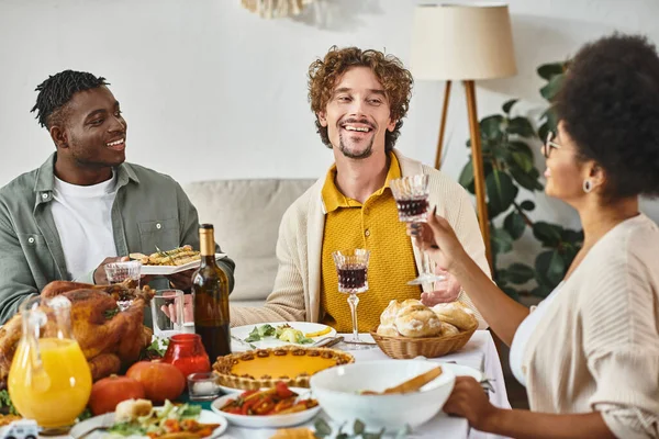 Feliz Día de Acción de Gracias, alegres amigos multiétnicos charlando en la mesa de banquetes con pavo asado - foto de stock