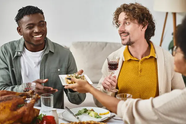 Tradición de acción de gracias, hombre americano africano alegre pasando comida a la hermana cerca de amigo feliz - foto de stock