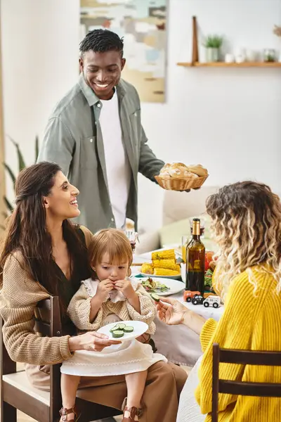 Happy Thanksgiving, curly woman feeding toddler daughter with cucumber near multiethnic friends — Stock Photo
