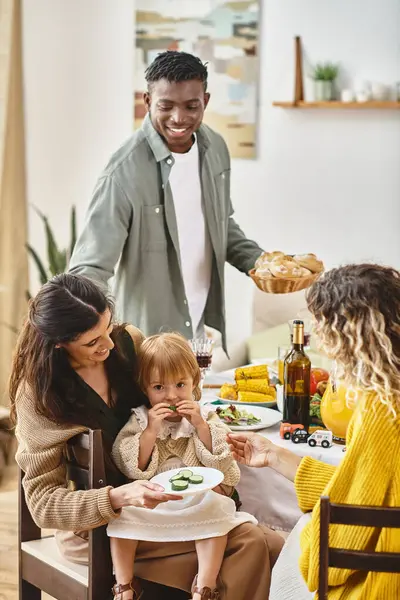 Feliz Día de Acción de Gracias, dos mujeres alimentando a un niño pequeño con pepino cerca de un amigo afroamericano - foto de stock