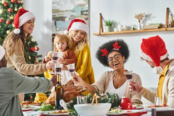 Gran familia alegre multiétnica tintineando sus gafas en la mesa festiva con sombreros de Santa Claus, Navidad - foto de stock