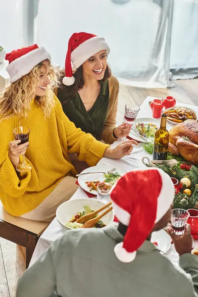 Alegre lgbt pareja en Santa sombreros sonriendo y celebrando la Navidad con sus parientes multiétnicos - foto de stock
