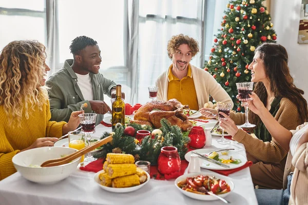 Alegres parientes multiétnicos tomando alegremente y disfrutando de la fiesta de Navidad con pavo y vino - foto de stock