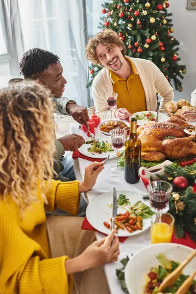 Alegres parientes multirraciales comiendo almuerzo festivo hablando activamente con el telón de fondo del árbol de Navidad - foto de stock