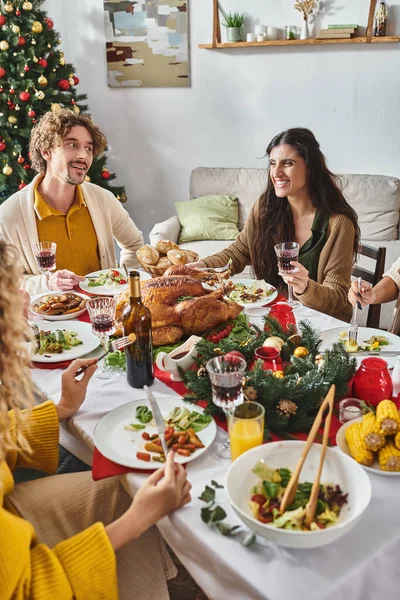 Обрезанный вид многорасовая семья наслаждаясь праздничным обедом и улыбаясь с елкой на заднем плане — стоковое фото