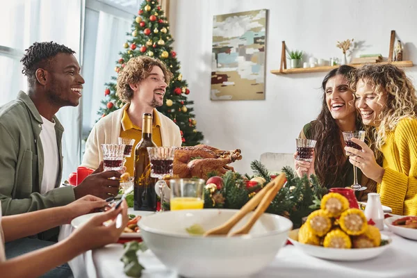 Alegre familia multiétnica disfrutando de fiesta con pavo y vino con fondo de árbol de Navidad - foto de stock