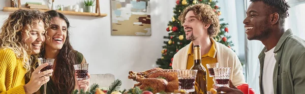Heureuse famille multiraciale riant et savourant le déjeuner avec dinde et vin, Noël, bannière — Photo de stock