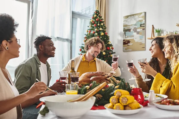 Grande famille multiethnique manger et claquer leurs verres à vin avec arbre de Noël sur fond — Photo de stock