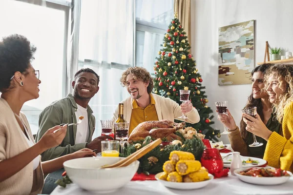 Familia multiétnica alegre pasar un buen rato comiendo almuerzo festivo con copas de vino levantadas, Navidad - foto de stock