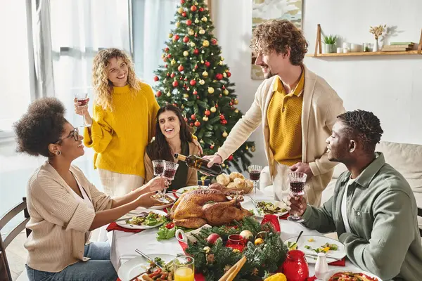 Gran familia multiétnica en trajes casuales disfrutando del almuerzo de Navidad y vertiendo un poco de vino en una copa - foto de stock