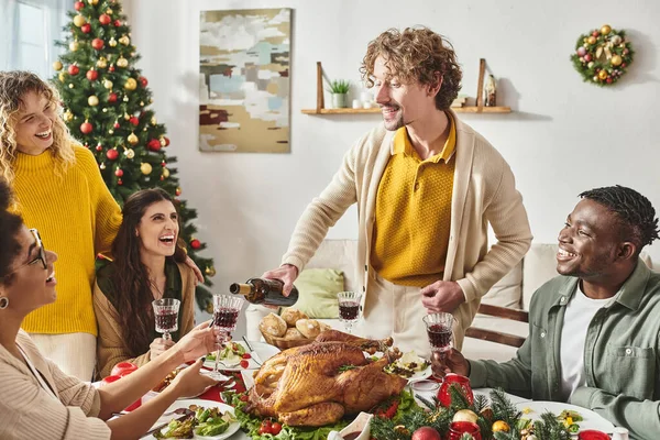 Giovane uomo sorridente circondato dai suoi parenti versando del vino in un bicchiere vicino all'albero di Natale — Foto stock