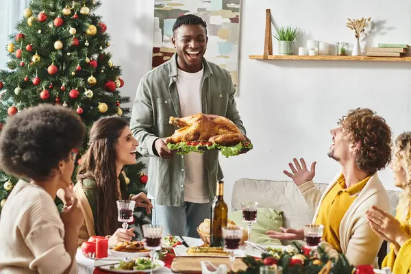 Alegre afroamericano rodeado por su familia multicultural sosteniendo plato con pavo, Navidad - foto de stock