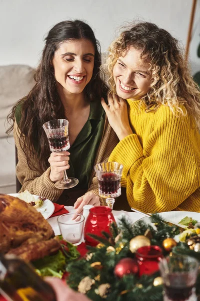 Alegre lgbt pareja abrazándose unos a otros y sonriendo sinceramente, sosteniendo copas de vino, Navidad - foto de stock