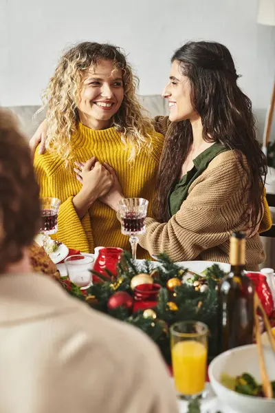 Dulce lgbt pareja cogido de la mano y mirando alegremente el uno al otro sentado en la mesa de Navidad - foto de stock