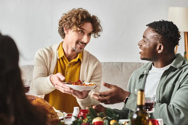 Jovens parentes multiculturais sentados à mesa de férias e compartilhando comida uns com os outros, Natal — Fotografia de Stock
