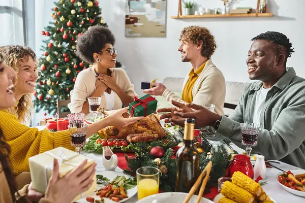 Big multicultural family sitting at festive table exchanging gifts with Christmas tree on backdrop — Stock Photo