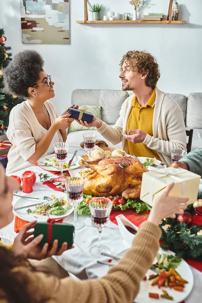 Jeunes membres de la famille multiethnique joyeux assis à la table de vacances et d'échanger des cadeaux, Noël — Photo de stock