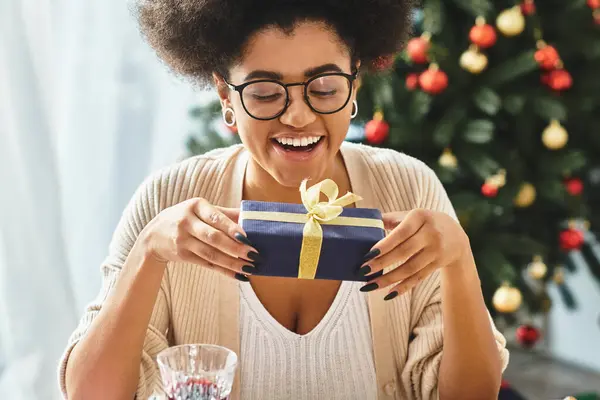 Joyous african american woman looking at her gift smiling cheerfully with Christmas tree on backdrop — Stock Photo