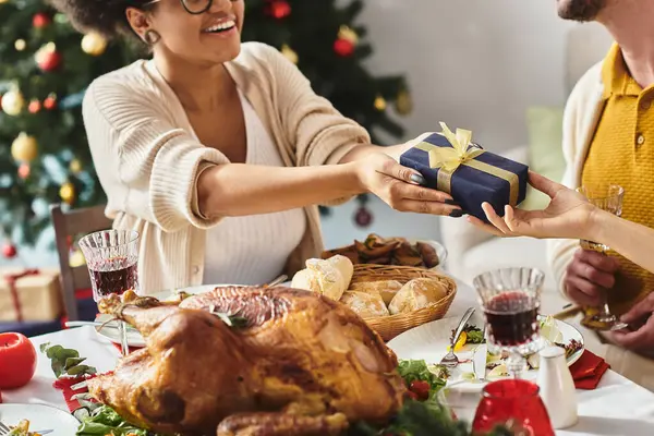 Cropped view of young cheerful african american woman giving Christmas gift sitting at holiday table — Stock Photo