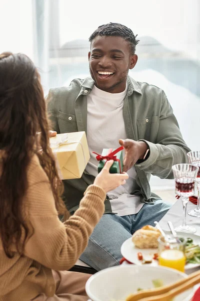Dois jovens parentes multirraciais rindo alegremente e trocando presentes na mesa festiva, Natal — Fotografia de Stock