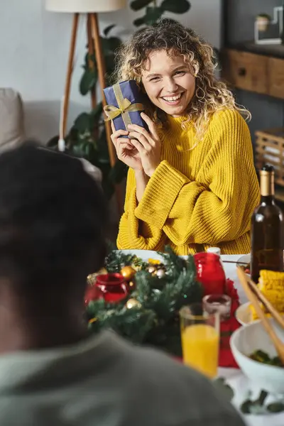 Heureuse femme blonde tenant cadeau de son parent et souriant joyeusement au déjeuner festif, Noël — Photo de stock