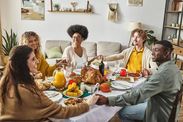 Família multiétnica feliz sentado à mesa festiva orando e de mãos dadas alegremente, Ação de Graças — Fotografia de Stock