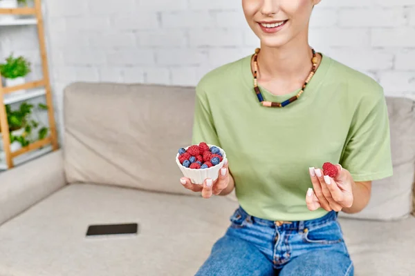 Vista cortada de mulher vegetariana com tigela de framboesas frescas e mirtilos na sala de estar — Fotografia de Stock