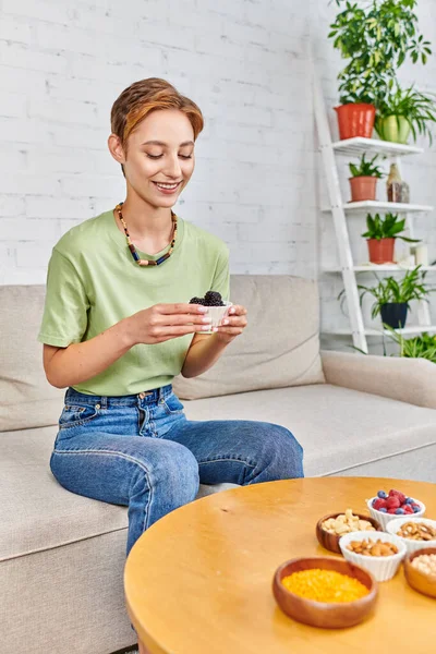 Mulher vegetariana alegre com amoras maduras perto do conjunto de alimentos à base de plantas na mesa na sala de estar — Fotografia de Stock