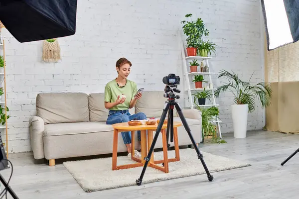 Blogueur vidéo avec smartphone près de la nourriture végétarienne et appareil photo numérique avec lampe softbox à la maison — Photo de stock