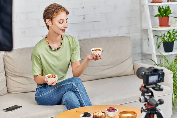 Молода вегетаріанка тримає миски з горіхами на дивані біля цифрової камери під час відеоблогів — стокове фото