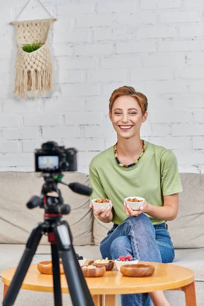 Mulher vegetariana alegre segurando tigelas com nozes perto de produtos à base de plantas e câmera digital em casa — Fotografia de Stock