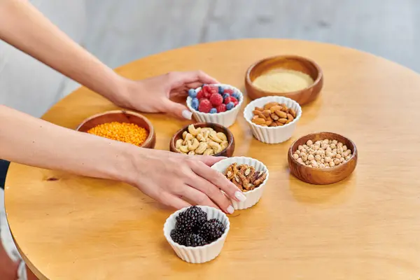 Mujer vegetariana colocar cuencos con bayas frescas y varios frutos secos con legumbres en la mesa en casa - foto de stock