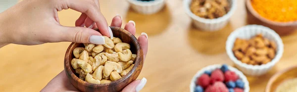 Partial view of woman with bowl of cashews above various plant-based food on blurred table, banner — Stock Photo