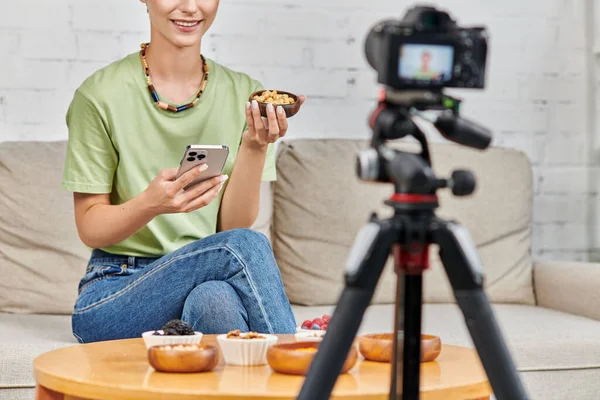 Donna ritagliata con smartphone e ciotola di anacardi vicino cibo vegetariano durante il video blog a casa — Foto stock