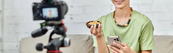Vista recortada de la mujer vegetariana con teléfono inteligente y tazón de anacardos cerca de la cámara digital, pancarta - foto de stock