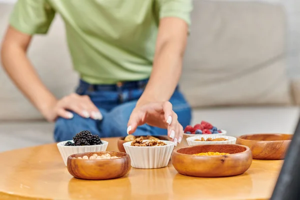Обрезанная размытая женщина берет чашу грецких орехов рядом со свежими ягодами и бобовыми на столе, вегетарианство — стоковое фото
