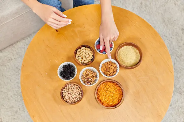 Обрезанная женщина указывает на стол с различными ягодами и орехами с бобовыми, растительное питание — стоковое фото